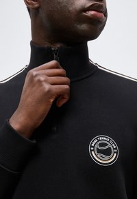 Το χέρι ενός άνδρα τραβάει το φερμουάρ ενός μαύρου τοπ με ψηλό γιακά και λογότυπο "Boss Tennis Club" στο στήθος.