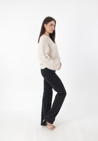 Maglione beige testurizzato abbinato a jeans neri a palazzo e sandali beige con punta aperta, posato su uno sfondo bianco.