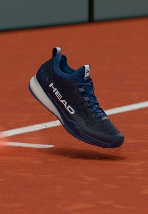 Blauwe sportieve schoen met een gestructureerd mesh bovenwerk, witte merkopdruk en een gedempte zool, zichtbaar in de lucht boven een gravelbaan.