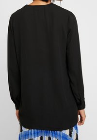 Black long-sleeve shirt with a rounded neck, smooth fabric, gathered shoulders, and buttoned cuffs, worn over a patterned skirt.