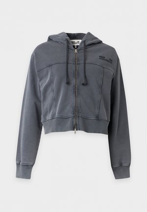 Vrouw draagt een donkergrijze cropped zip-up hoodie, zwarte top, wijdvallende blauwe spijkerbroek en zwarte puntige platte schoenen met zilveren gespdetails.