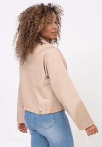Veste en cuir beige avec un col structuré, des panneaux latéraux et des détails de couture visibles, associée à un jean en denim bleu clair.