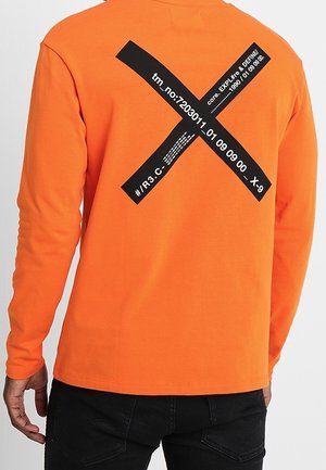 Langärmliges Shirt aus leuchtend orangefarbenem Baumwollstoff mit einem kühnen schwarzen X-förmigen Grafik- und Textdesign auf der Rückseite. Schlichte Vorderseite, weiche Textur.