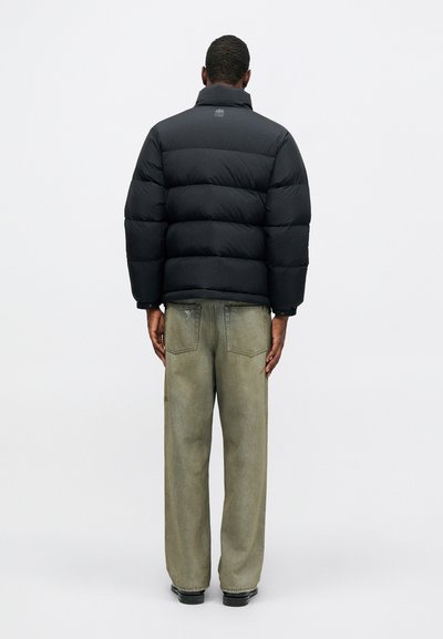 Vyras stovi atsukęs nugarą, vilkėdamas juodą pūkinę striukę ir laisvas, blukusias žalias džinsus, prieš paprastą baltą foną.