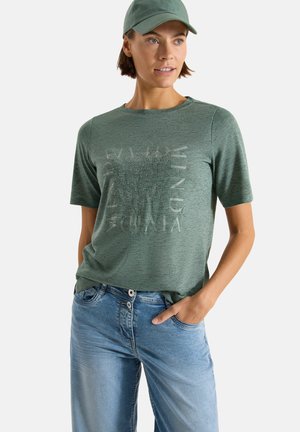 Kvinde iført en grøn kasket, grøn T-shirt med korte ærmer og subtil tekst samt lyseblå jeans, står med den ene hånd i lommen.