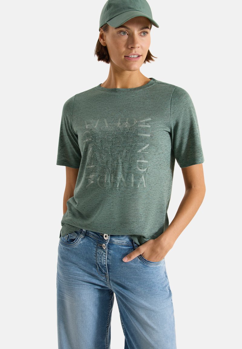 Femme portant une casquette verte, un t-shirt vert à manches courtes avec un texte discret, et un jean bleu clair, debout avec une main dans la poche.