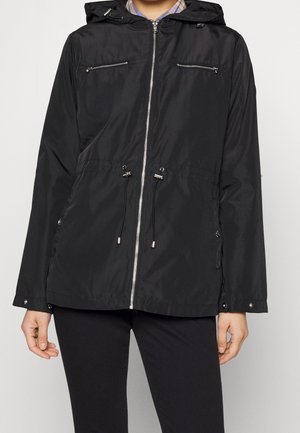 Henkilö, jolla on musta hupullinen takki, jossa on etuvetoketju, kaksi vetoketjullista rintataskua ja säädettävät kiristysnauhat vyötäröllä.