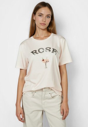 Frau trägt ein blassrosa T-Shirt mit dem Aufdruck "ROSE all day" und einer Pailletten-Grafik eines Cocktailglases, kombiniert mit hellbeigen Hosen.