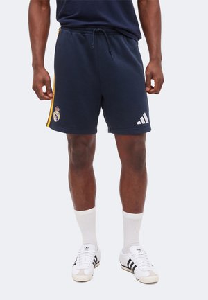 Bărbat purtând pantaloni scurți albastru închis Real Madrid cu dungi galbene, logo alb Adidas, șosete albe și adidași albi Adidas, stând drept.
