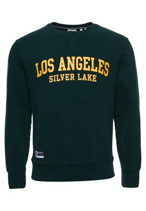 Mężczyzna w zielonej bluzie "LOS ANGELES SILVER LAKE", niebieskich podwiniętych dżinsach, białych trampkach, srebrnym łańcuszku na szyi i bransoletce na tle białej ceglanej ściany.