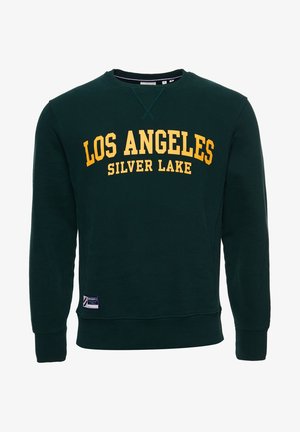 Mand iført grøn "LOS ANGELES SILVER LAKE" sweatshirt, blå opkørte jeans, hvide sneakers, sølvkæde og armbånd foran en hvid murstensvæg.