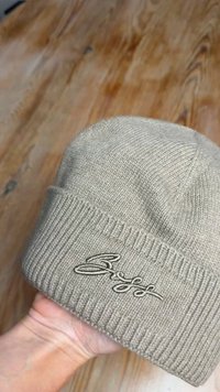 Kötött bézs sapka hajtott bordázott mandzsettával. Az elején világosbarna hímzett logóval, sima textúrával.