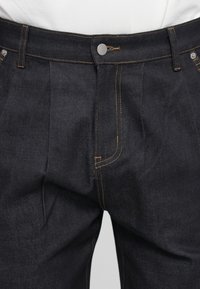 Donkerdenim jeans met een aansluitend ontwerp, voorzien van een knoopsluiting en gouden stiksels. Plooien voegen textuur toe aan de stof.