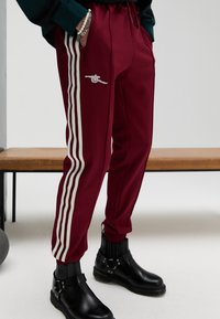 Tumši sarkanas sporta bikses ar baltām sānu svītrām, elastīgu vidukli un kabatām ar rāvējslēdzēju. Tās ir ar mazu izšūtu dizainu un šaurām aprocēm.