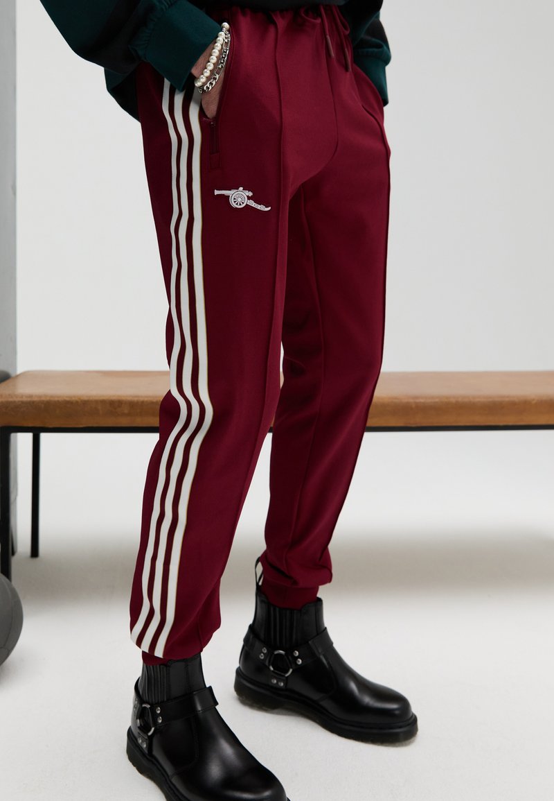 Vinröda träningsbyxor med vita sidoremsor, elastisk midja och blixtlåsförsedda fickor. Har en liten broderad design och avsmalnande muddar.