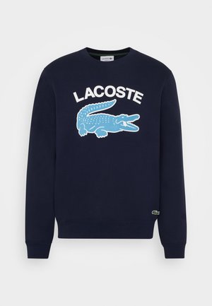 Mężczyzna w granatowej bluzie Lacoste z niebieskim logo krokodyla, kremowych spodniach i białych sneakersach z czerwonymi i granatowymi paskami.