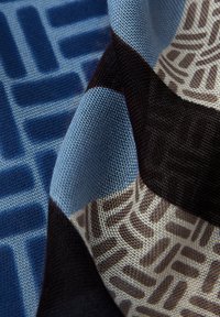 Tekstilstof med et geometrisk mønster af sammenlåste former i blå, brune og beige toner, der fremviser en tekstureret vævning.