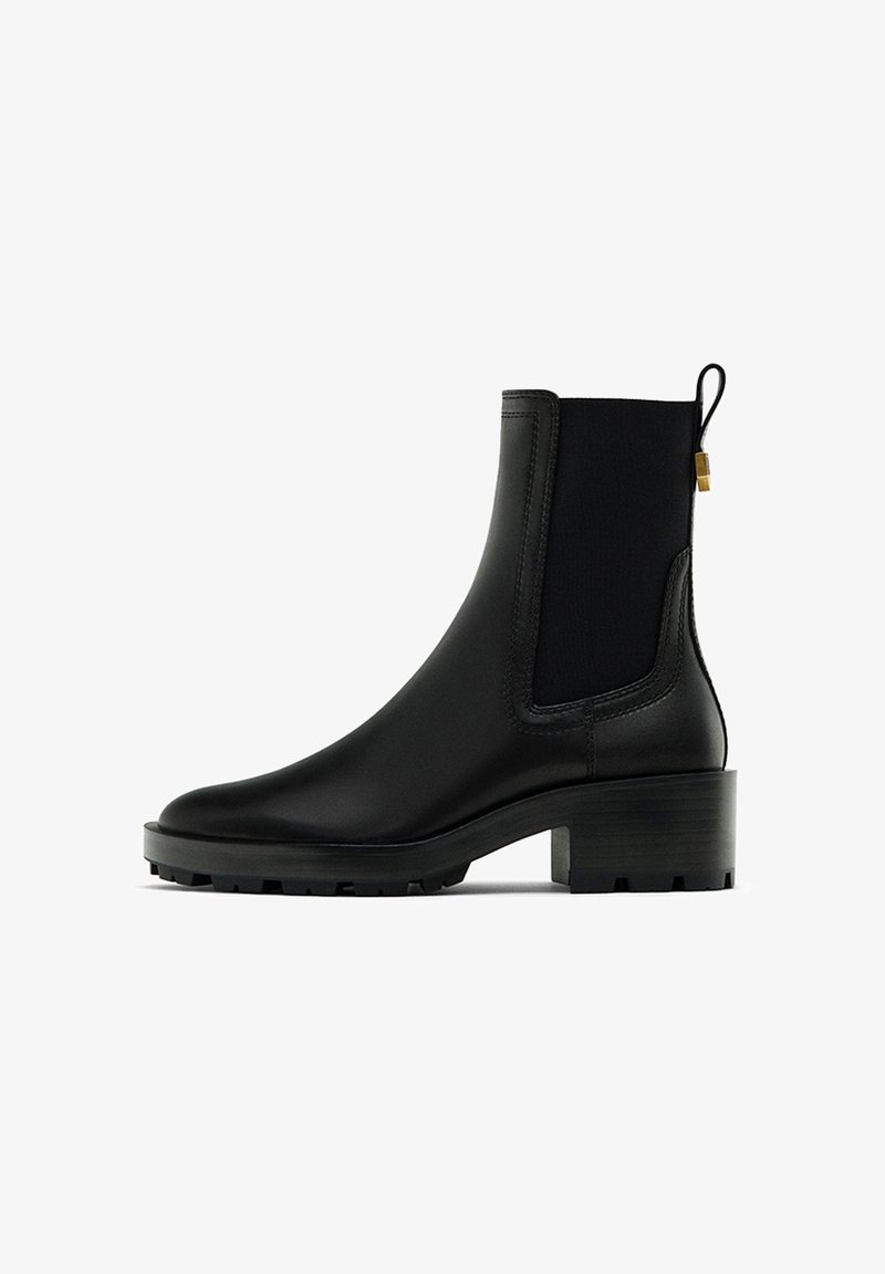 Bottine Chelsea en cuir noir avec talon épais, panneau latéral élastique, languette avec accent doré et semelle à relief robuste.