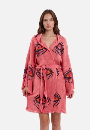 Vrouw draagt een losse roze badjas met riem, versierd met grote geborduurde ogen in oranje, blauw en zwart op de mouwen en het lichaam.