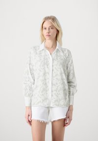 White long-sleeve shirt with a subtle floral pattern, buttoned down front, and a collar. Paired with white frayed shorts.