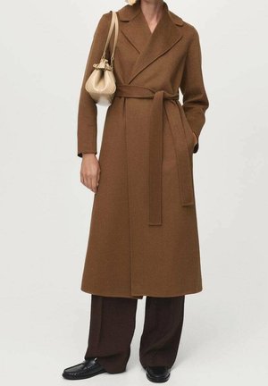 Persoon draagt een lange bruine jas met ceintuur over een donkere broek, met een kleine beige handtas over de schouder, staand tegen een effen achtergrond.