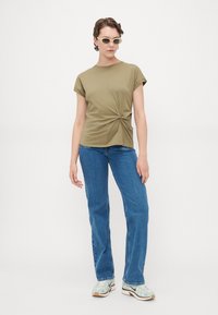 Olivengrønn t-skjorte med vridd frontdesign, korte ermer, kombinert med blå jeans og lyse joggesko. Jevn tekstur på stoffet.