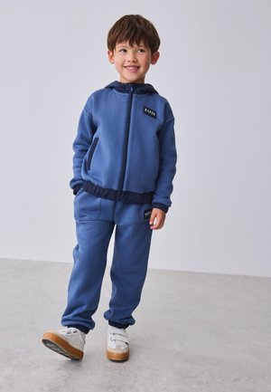 Naeratav noor poiss seisab siseruumides, kandes sinist lukuga kapuutsiga dressipluusi ja sobivaid dressipükse valgete spordijalatsitega.