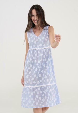 Mujer sonriente con cabello largo y oscuro, vistiendo un vestido sin mangas azul claro con un patrón floral blanco, posando frente a un fondo claro y liso.