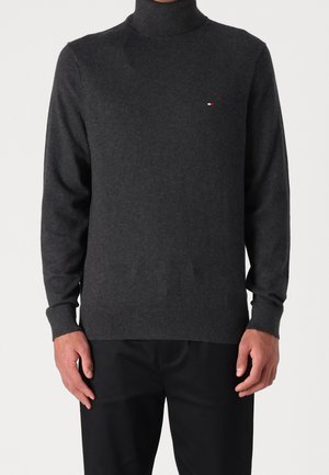 Mann in einem dunkelgrauen Rollkragenpullover mit einem kleinen rechteckigen Logo auf der linken Brust und schwarzen Hosen vor einem schlichten Hintergrund.