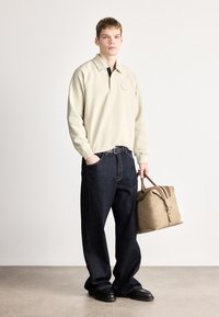 Bézs pulóver hímzett logóval, sötétkék bőszárú farmernadrág, valamint kontrasztos pántokkal és cipzárral ellátott barna bőr utazótáska.