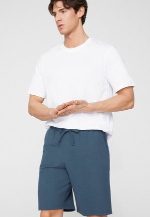 Młody mężczyzna w białej, gładkiej koszulce z krótkimi rękawami i niebieskich szortach z troczkiem, z dłońmi złączonymi na wysokości talii, patrzący w bok.