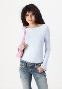 Light blue long-sleeve top with a boat neckline, stretchy fabric, and flared cuffs paired with dark denim jeans and a pink handbag.