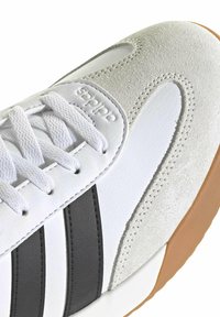 Primer plano de una zapatilla blanca con detalles en ante beige, franjas laterales negras, cordones blancos y suela de goma color caramelo.