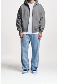 Sudadera gris con cremallera y bolsillos frontales, llevada sobre una camiseta blanca. Unos vaqueros sueltos de color azul claro y unas zapatillas grises y azules completan el conjunto.