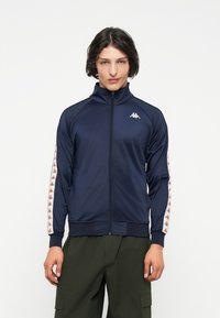 Tamno plava zip-up trenerka s visokim ovratnikom, s bijelim i crvenim logotipima na rukavima. Glatka tkanina i rebrasti manžeti.