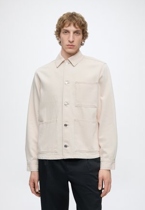 Jeune homme aux cheveux courts et bouclés porte une veste beige à boutons avec trois poches avant et un pantalon noir sur un fond uni.