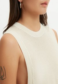 Närbild på en person som bär en ärmlös beige stickad topp med textur, silverfärgade ringörhängen och en synlig arm tatuering på vänster arm.