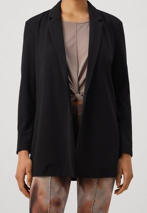 Personne portant un blazer noir ouvert sur un haut taupe noué et un legging marron et gris à motifs, debout devant un fond uni.