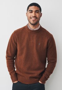 Brauner Strickpullover mit geripptem Kragen und Bündchen. Verfügt über ein kleines Logodetail auf der linken Brust. Wird über einem karierten Hemd getragen.