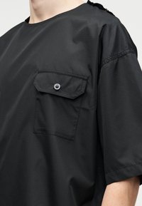 Chemise noire surdimensionnée à manches courtes avec une poche poitrine boutonnée et un détail en tissu sur l'épaule gauche, portée par une personne.