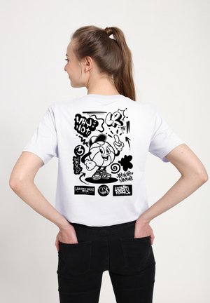 Młoda kobieta z kucykiem, ubrana w białą koszulkę z czarną postacią w stylu graffiti i napisem na plecach.