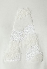 Hanorac alb cu un litera "A" texturată, decorată cu material pufoasă și paiete, creând un design tactil contrastant.
