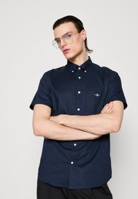 Young man with slicked-back hair and glasses, wearing a navy short-sleeve button-up shirt with arms crossed against a plain background.