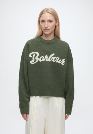 Kobieta o prostych blond włosach, ubrana w za duży zielony sweter z napisem "Barbour" oraz białe spodnie o szerokich nogawkach na jednolitym tle.