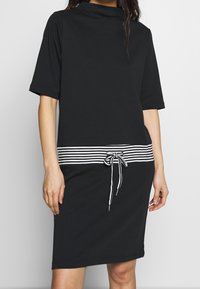 Vestido negro con cuello de embudo, mangas cortas y una cinturilla con corbata a rayas. La tela parece suave y transpirable.