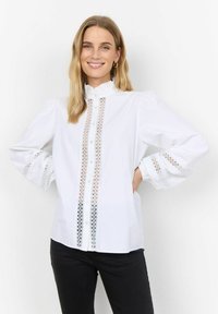 Camicia bianca con colletto arricciato, dettagli di pizzo lungo il centro e accenti in pizzo sulle maniche a sbuffo. Chiusura con bottoni visibile.