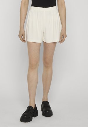Vrouw draagt losse witte shorts, zwart topje en zwarte stevige loafers, staand tegen een effen achtergrond.