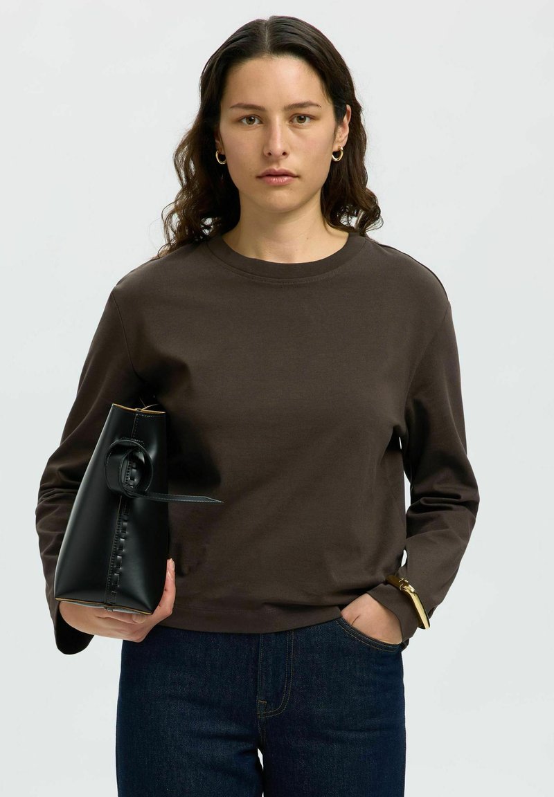 Smeđa dukserica dugih rukava s okruglim izrezom, uparena s tamnim trapericama. Model drži elegantnu crnu torbu s okruglim detaljem.