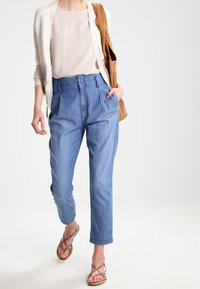 Lehká béžová blůza, krémový cardigan, džíny s vysokým pasem v modré barvě, hnědé ploché sandály a hnědá kabelka. Neformální outfit.