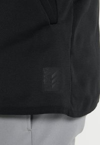 Veste en maille noire avec un tissu texturé, une poche zippée et une étiquette logo noire avec trois bandes horizontales sur le côté inférieur.